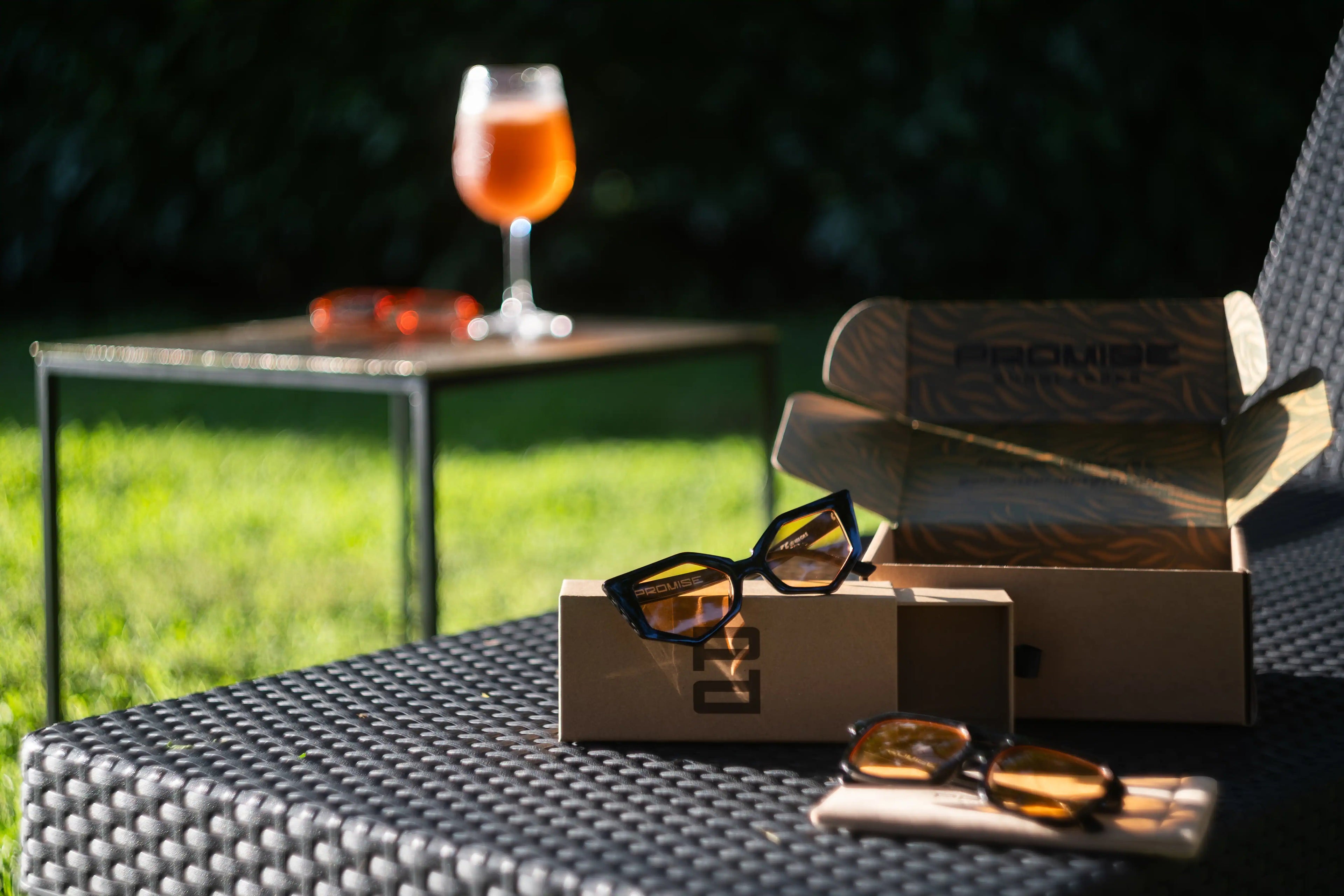 Occhiali da sole PROMISE con lenti ambrate su lettino in giardino, packaging ecologico aperto e mood aperitivo.